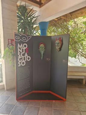 Un pannello espositivo grigio con bordo rosso, montato sotto la pergola di un villaggio turistico: porta la scritta Montescaglioso e due maschere di cartapesta appese, una verde con la barba lunga e una rossa con le corna.