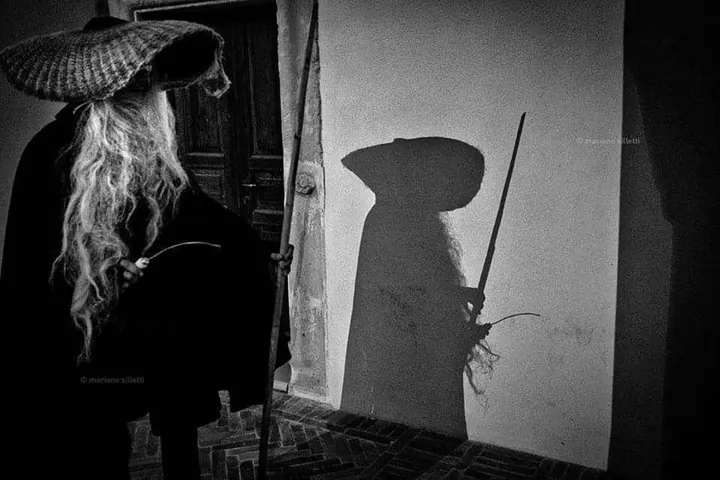 Un Cucibocca in bianco e nero, con il cappello a falda larga e il bastone; la sua ombra è proiettata sul muro