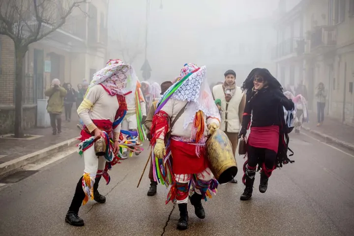 Maschere de L’Màshkr in corteo nella nebbia lungo una via di Tricarico, una di loro porta un campanaccio